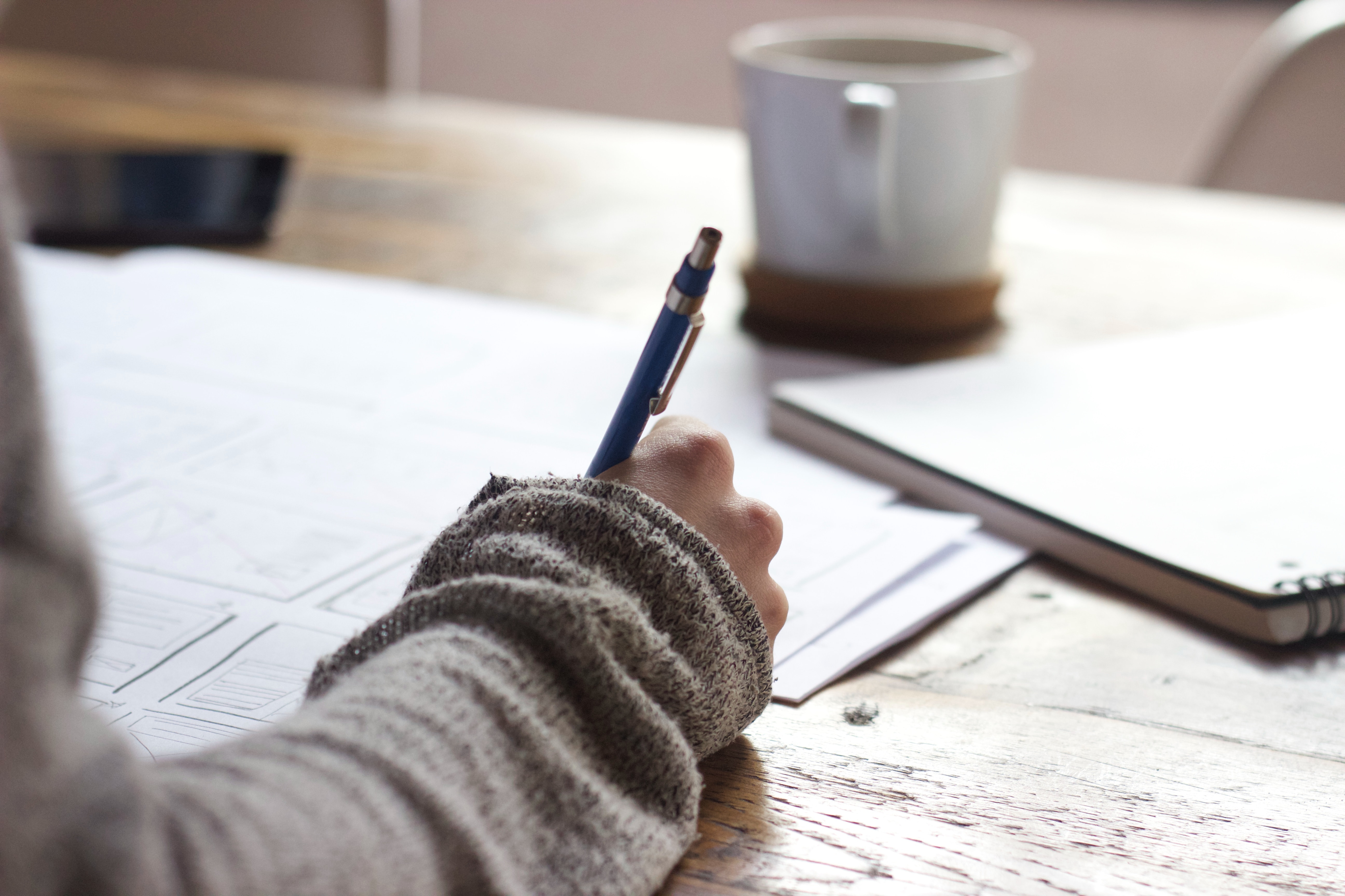 person writing on a brown wooden table with white ceramic mug